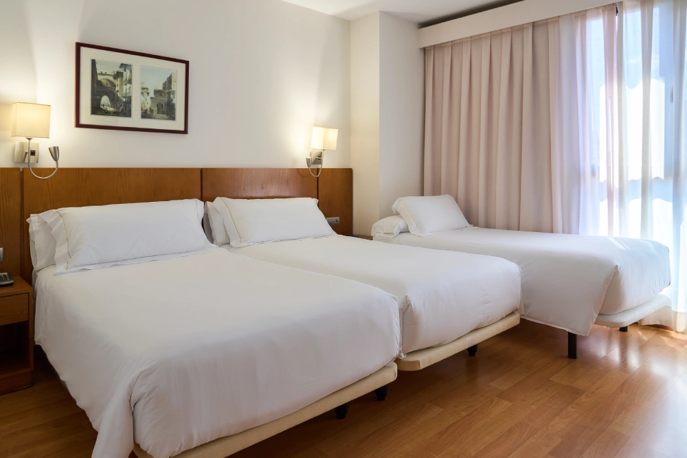 Habitaci&oacute;n de hotel con dos camas gemelas y cortinas beige junto a una ventana