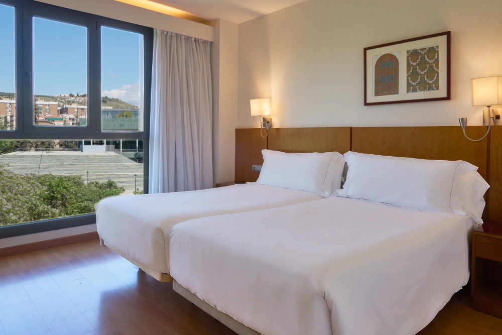 Dormitorio con cama doble blanca, l&aacute;mparas y grandes ventanales.
