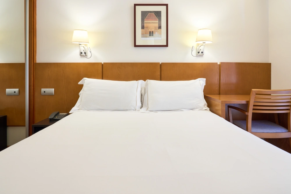 Cama doble con almohadas blancas y cabecero de madera en habitaci&oacute;n de hotel
