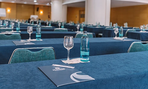 Sala de conferencias preparada con mesas, copas y botellas de agua.