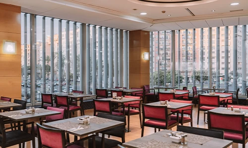 Restaurante elegante con sillas rojas y mesas vac&iacute;as cerca de grandes ventanas.