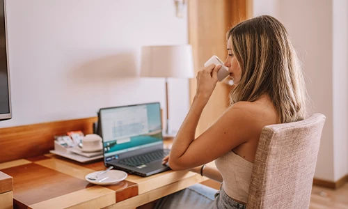Mujer trabajando en una laptop y tomando un caf&eacute; en una habitaci&oacute;n.