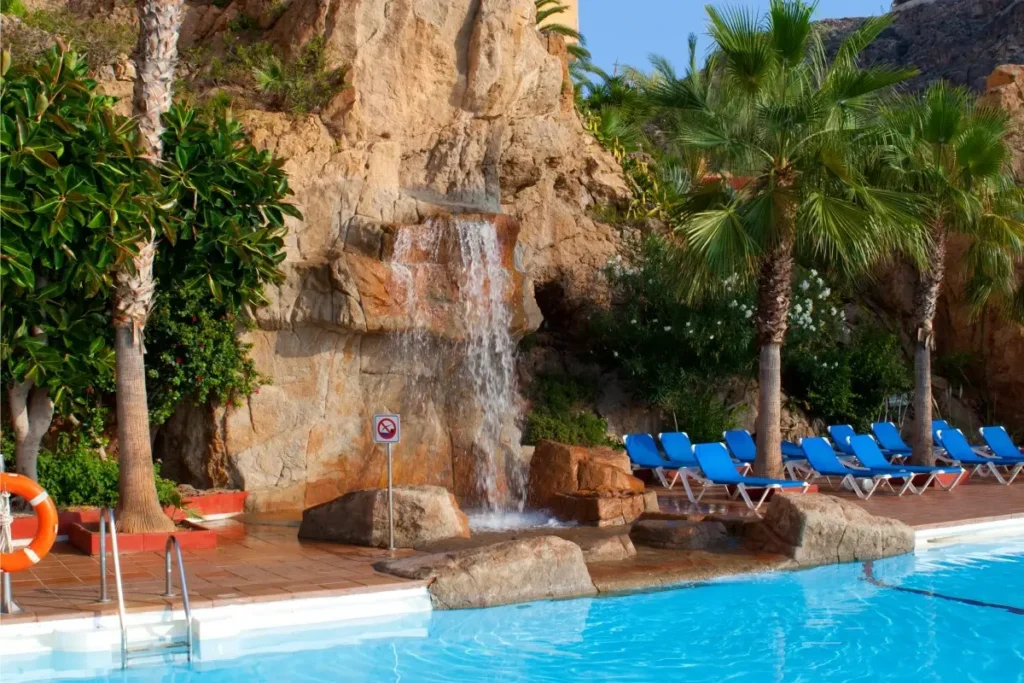 Cascada sobre rocas, piscina y tumbonas azules entre palmeras en un hotel