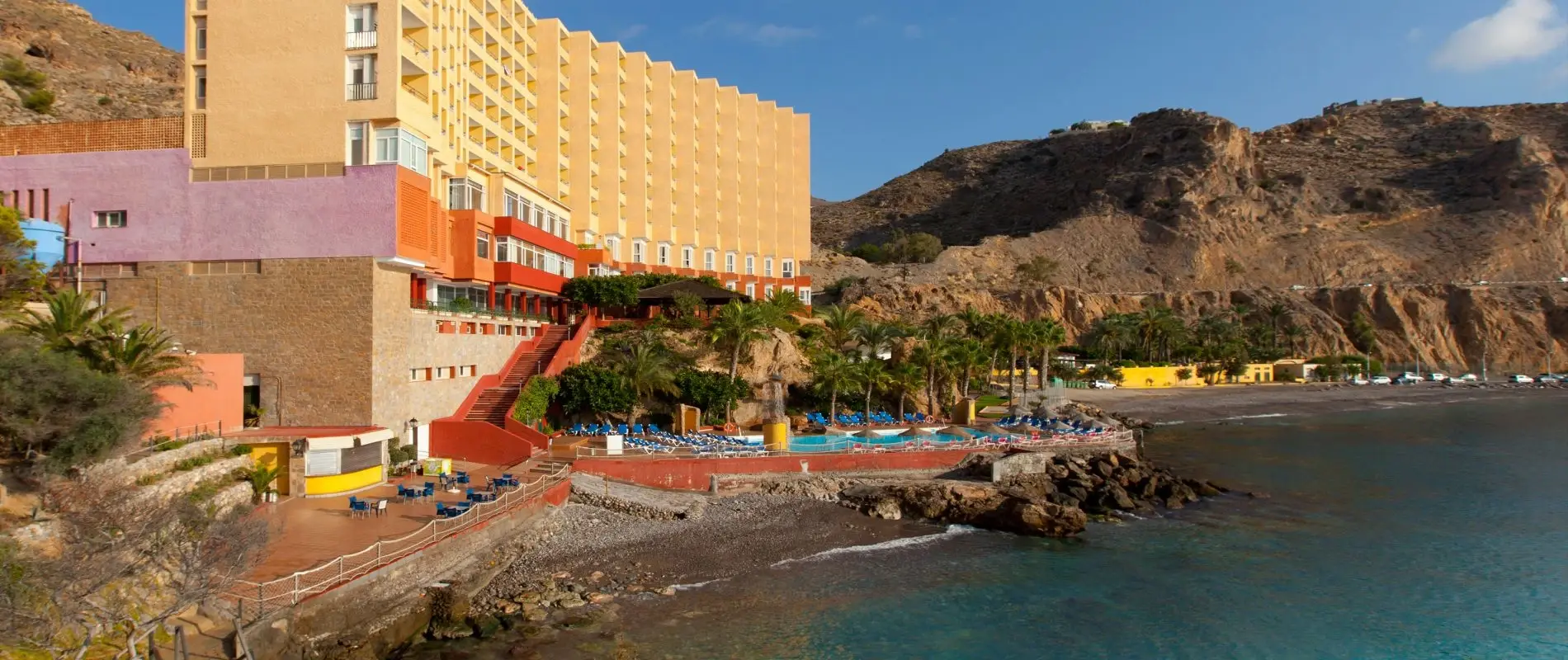 Hotel junto al mar, con piscina y palmeras frente a monta&ntilde;as rocosas.