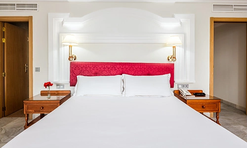 Cama de hotel con cabecero rojo, l&aacute;mparas y mesas laterales con tel&eacute;fonos y flores.