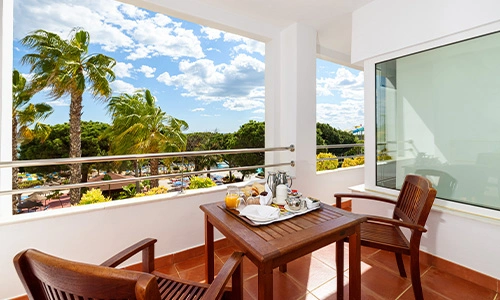 Balc&oacute;n soleado con mesa, sillas y desayuno, vista a palmeras y cielo azul.