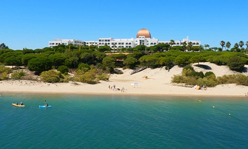Playa con kayaks y edificio blanco con c&uacute;pula al fondo bajo cielo despejado.