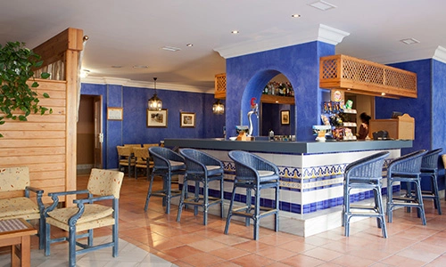 Bar con barra azul, sillas altas y mesas en un espacio decorado en tonos c&aacute;lidos.