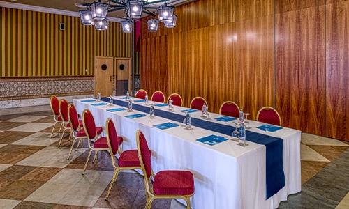 Sala de conferencias con mesa en U, sillas rojas y papeles sobre la mesa.