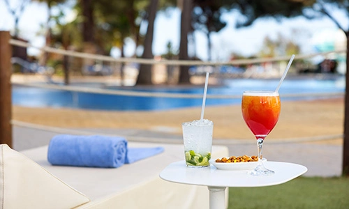 Bebidas junto a una piscina, con una toalla y &aacute;rboles de fondo.