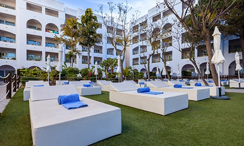 Terraza con camastros blancos y toallas azules frente a edificio blanco.