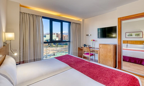 Habitaci&oacute;n de hotel moderna con cama doble, ventana grande y TV en la pared.