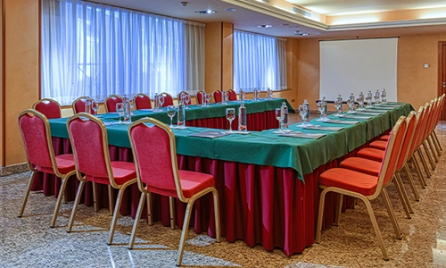 Sala de conferencias con mesa en U, sillas rojas y copas de agua.