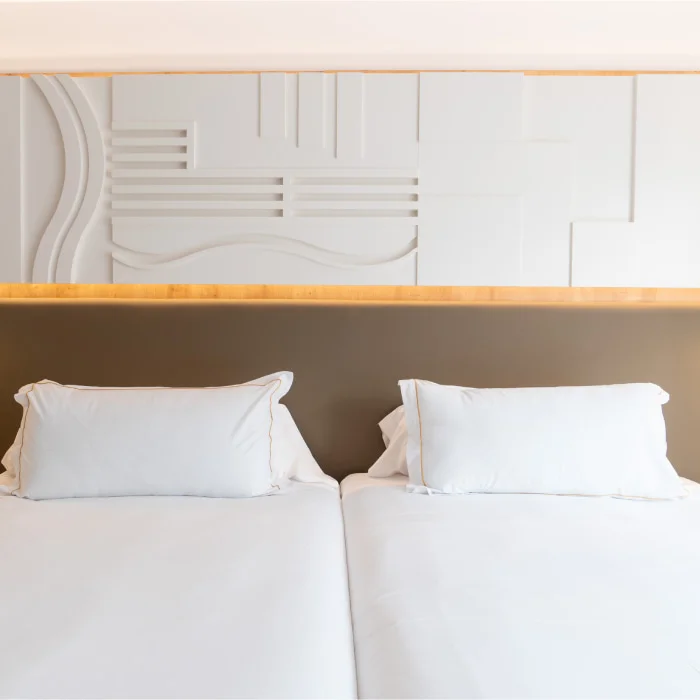 Cama con s&aacute;banas y almohadas blancas, vista desde cabecero y pared blanca.