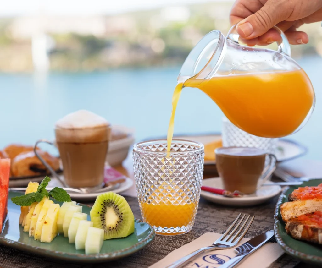Mano sirviendo t&eacute; naranja en vaso de vidrio sobre mesa con frutas y pasteles
