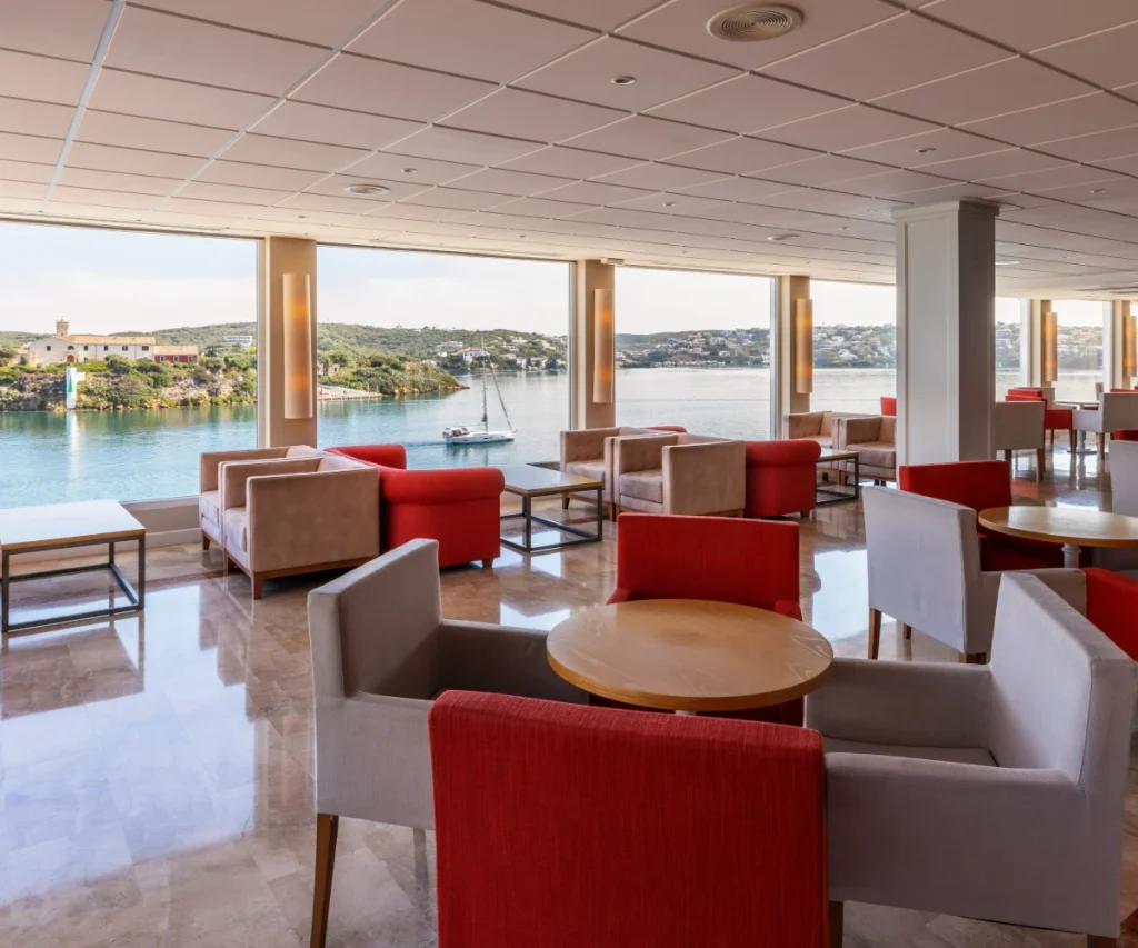 Sala moderna con sillones rojos, mesas y vista al lago desde un porche.