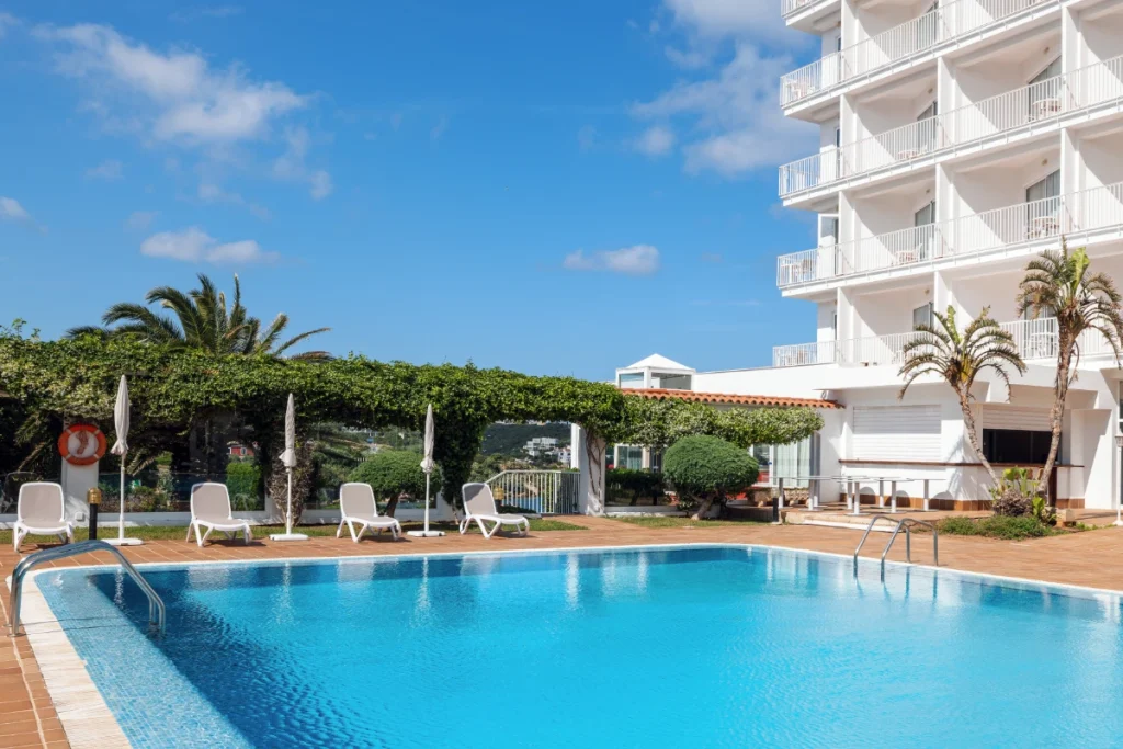 Piscina azul con tumbonas y edificio blanco, palmeras y cielo despejado