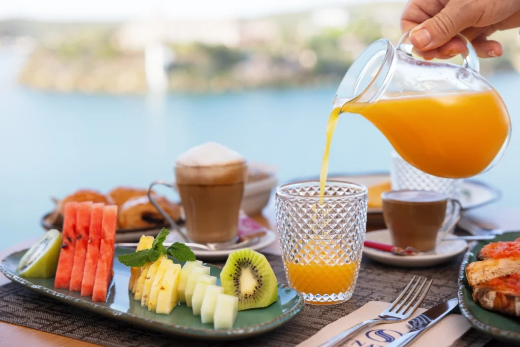 Sirviendo jugo naranja en vaso de cristal, con frutas y desayunos al fondo