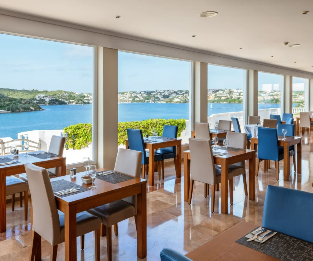 Comedor con mesas y sillas azules, vista al mar y ventanales.