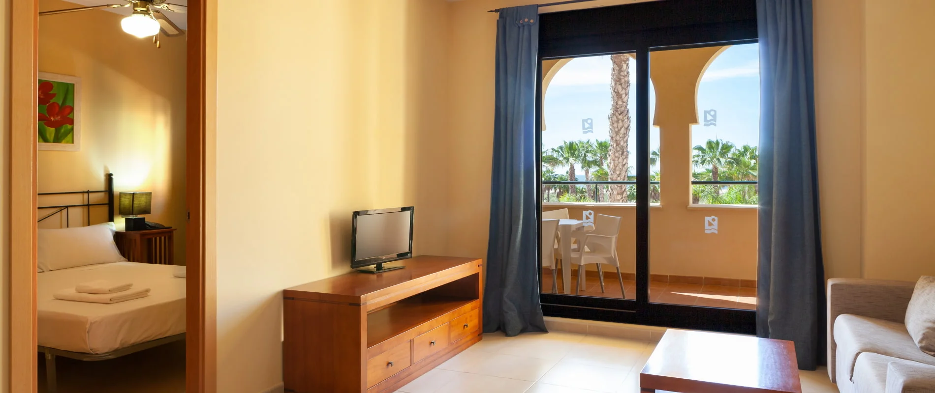 Interior de habitaci&oacute;n de hotel con cama, TV y cortinas azules, vista al balc&oacute;n