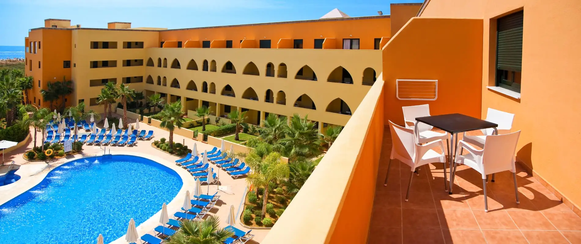 Hotel con piscina azul, jardines y terraza con mesa y sillas al atardecer
