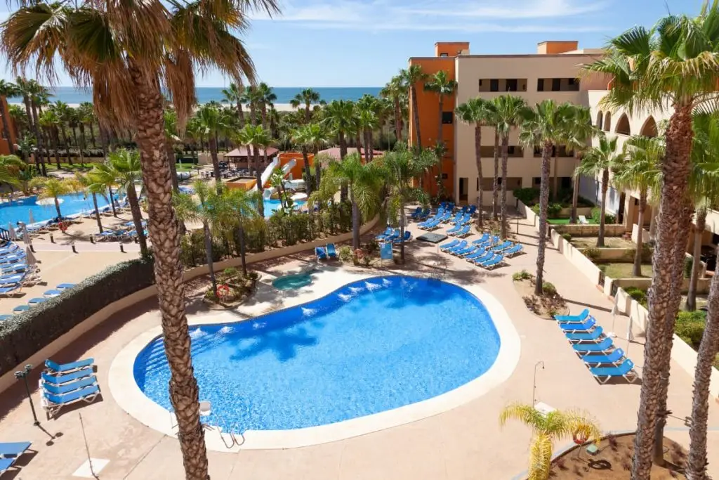 Hotel con piscina azul, tumbonas y palmeras junto al mar.