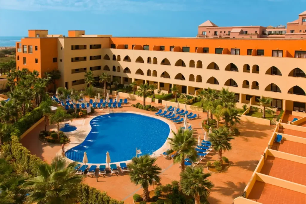 Piscina ovalada con tumbonas y edificio hotelero junto al mar