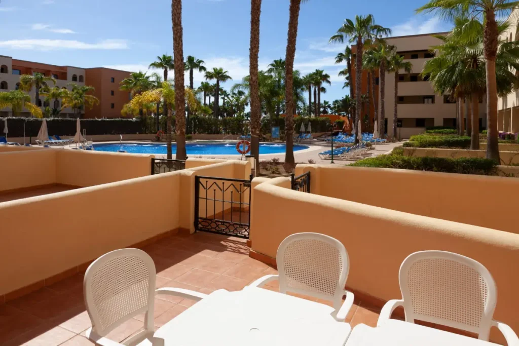 Terraza con sillas blancas y vista a piscina y palmeras en hotel.
