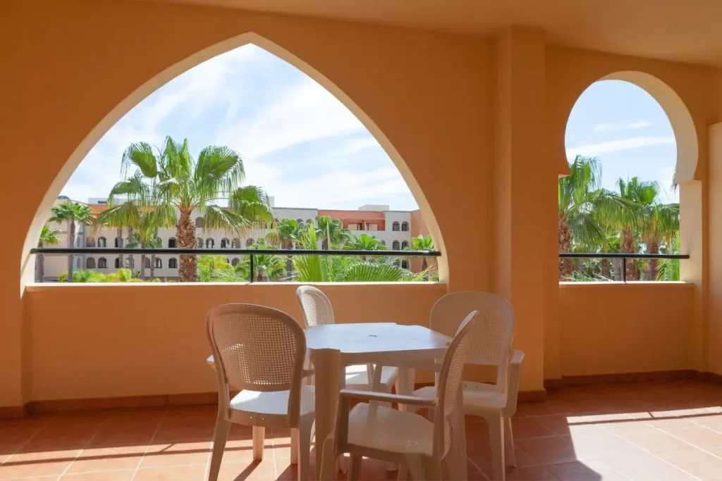 Terraza con mesa y sillas, ventanas arqueadas y vista a palmeras y hotel.