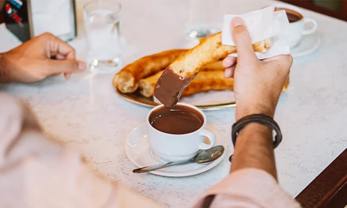Persona remojando un churro en chocolate caliente sobre una mesa de caf&eacute;.
