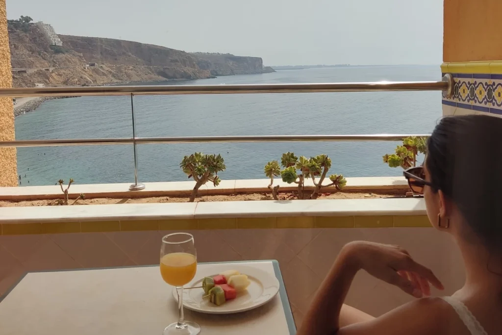 Mujer en balc&oacute;n frente al mar, mesa con bebida y fruta, acantilados al fondo