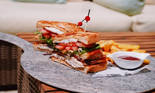 S&aacute;ndwich club con papas fritas y salsa en una mesa al aire libre.