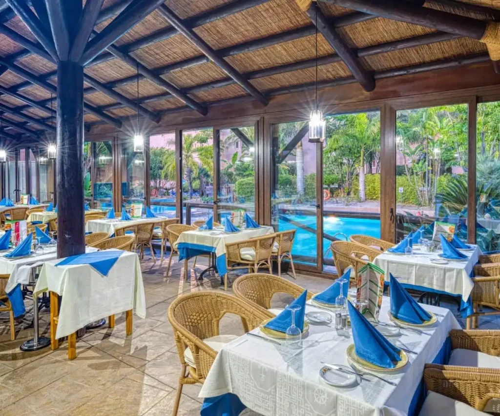 Comedor de restaurante con mesas con mantel blanco y azul, junto a piscina exterior