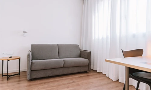 Sala minimalista con sof&aacute; gris, mesa lateral y cortinas blancas.