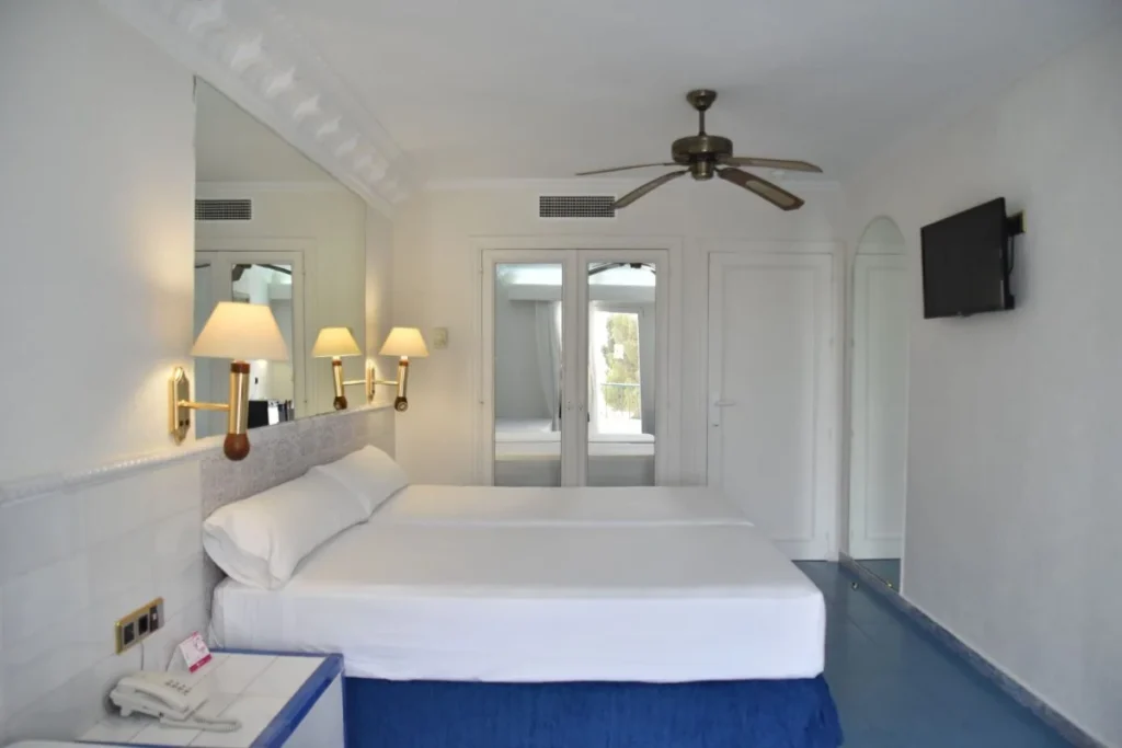 Habitaci&oacute;n de hotel blanca con cama, l&aacute;mparas y ventilador de techo