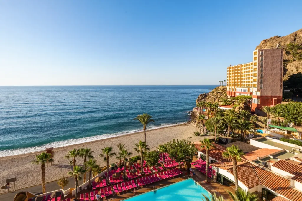 Hotel junto a la playa, con piscina y palmeras sobre el acantilado.