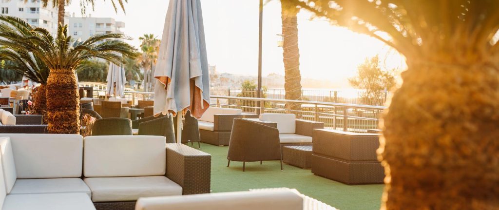 Terraza con muebles, sombrilla y palmeras al atardecer, con luz c&aacute;lida.