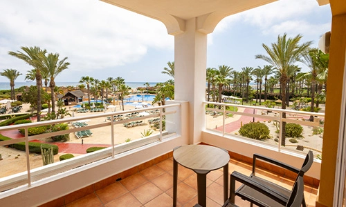 Balc&oacute;n con vistas a palmeras, piscinas y el mar en el horizonte.