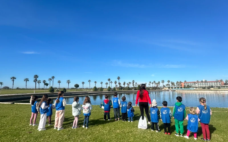 Ni&ntilde;os con camisetas y mochila en un lago, palmeras y cielo azul.