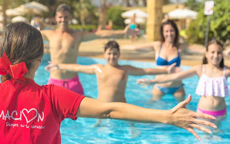 Ni&ntilde;os y adultos en una piscina, uno con camiseta roja se&ntilde;alando al agua.