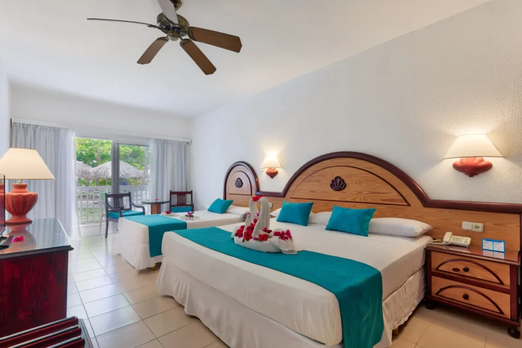 Habitaci&oacute;n de hotel con cama doble, almohadas azules y ventilador de techo