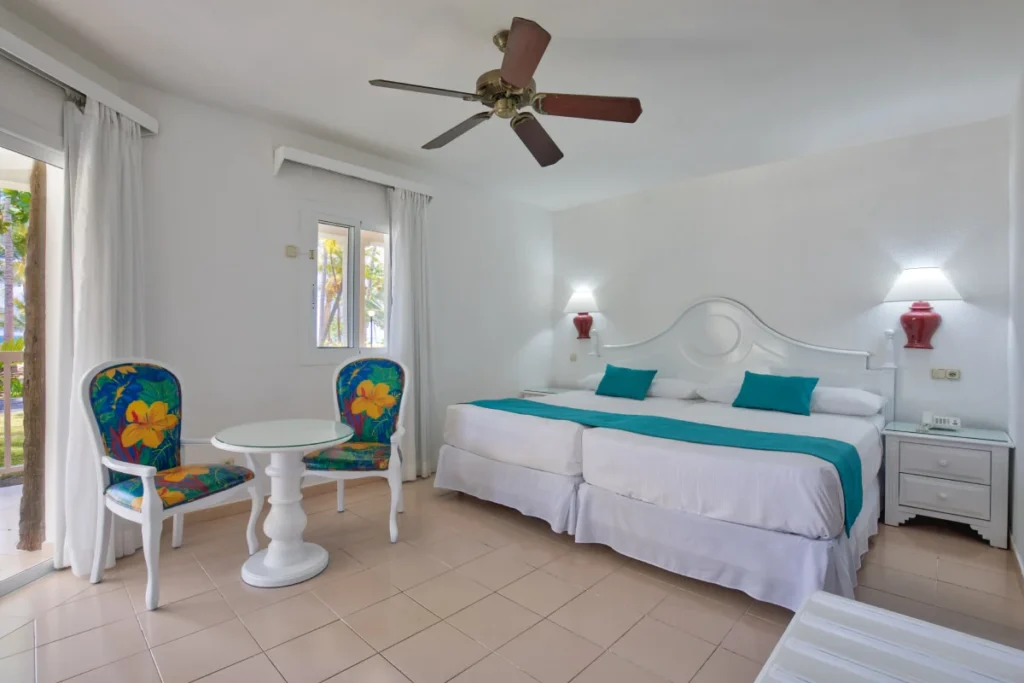 Dormitorio blanco con cama, l&aacute;mparas, sillones coloridos y ventilador de techo