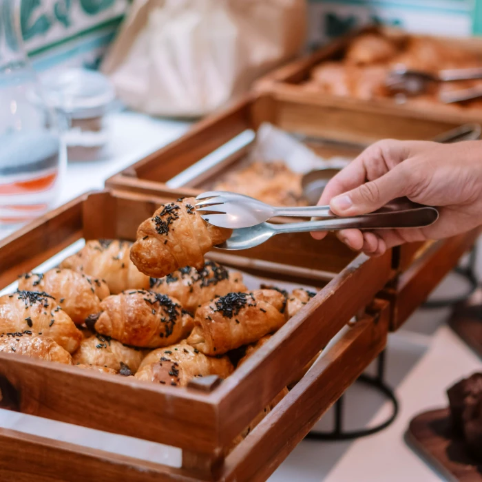 Mano tomando croissant con tenedor y cuchara en bandeja de madera.