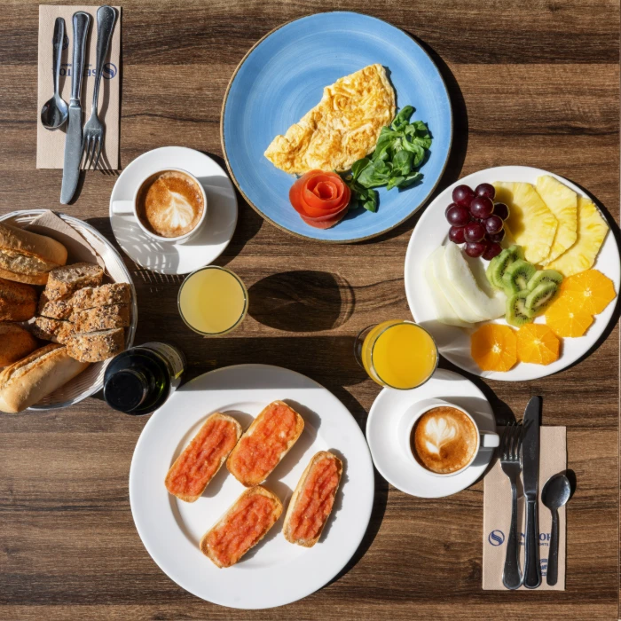 Desayuno con frutas, pan, caf&eacute;, jugo y tortilla en una mesa de madera.