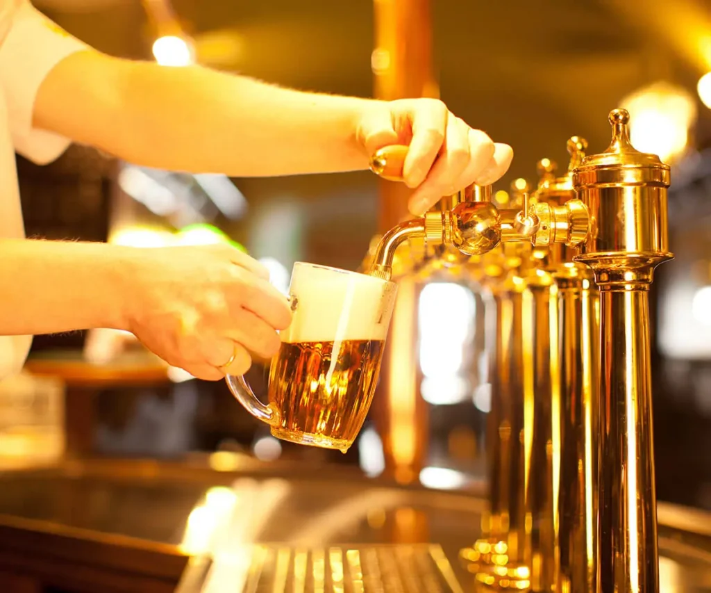 Manos sirven cerveza en jarra desde un barril con grifo dorado y luces c&aacute;lidas