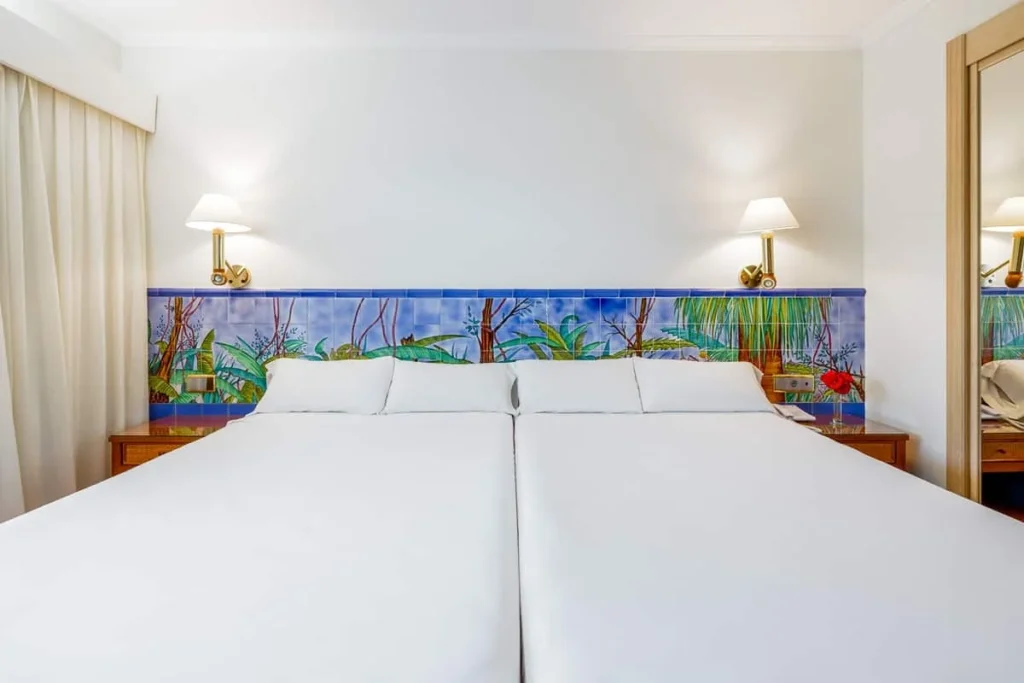 Habitaci&oacute;n de hotel con dos camas blancas y mural tropical en la pared