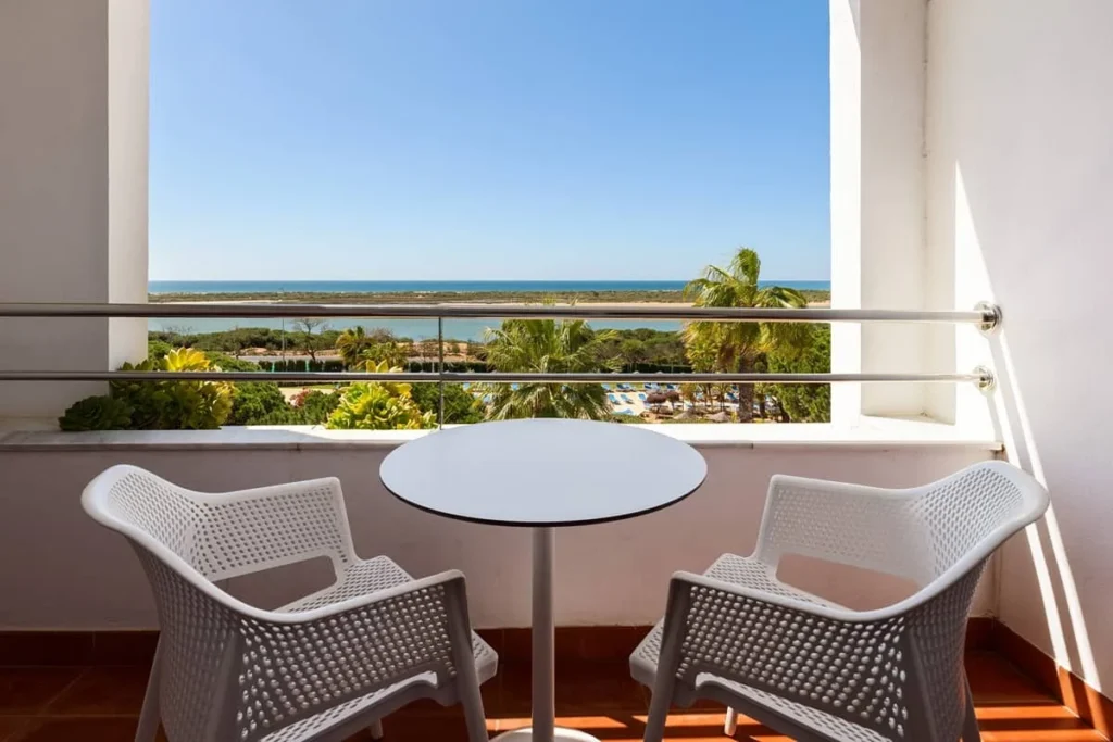 Balc&oacute;n con mesa y sillas blancas, vista al mar y palmeras al atardecer