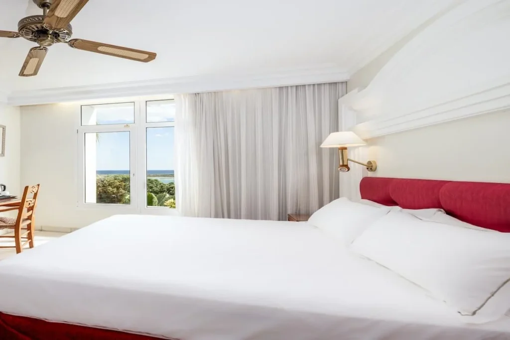 Dormitorio moderno con cama blanca, cabecero rojo, l&aacute;mpara y ventilador.