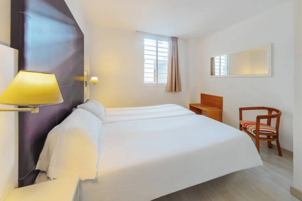 Dormitorio moderno con cama blanca, mesita, l&aacute;mparas y ventana con cortinas beige