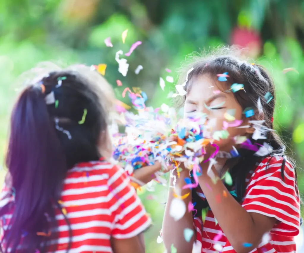 Ni&ntilde;as celebran al aire libre con confeti, sonriendo y con los ojos cerrados.